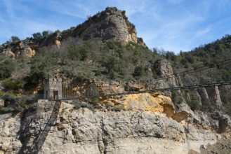 A rocky mountain landscape with dense vegetation under a bright blue sky, suspension bridge,