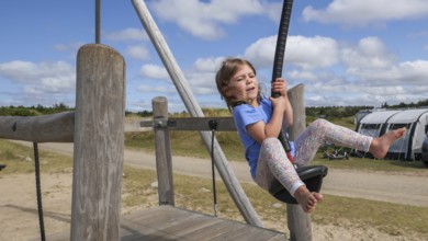 A child happily jumps on a zip wire in a wide, sunny playground setting with a cloudy, blue sky