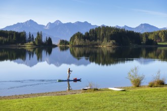 A person paddles with a surfboard on a calm lake with mountains and forest in the background,