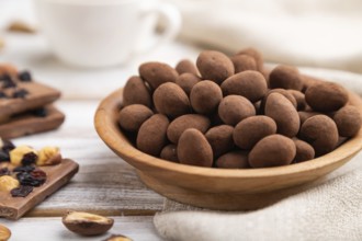 Almond in chocolate dragees in wooden plate and a cup of coffee on white wooden background and