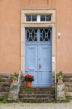 A blue front door with iron decorations on a pink façade, surrounded by potted plants,