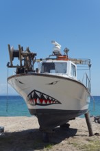 A small fishing boat with a painted shark face on the beach in front of a clear sky, shipyard,