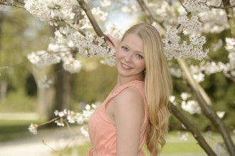 Woman with blonde hair in a pink dress smiles while leaning against a cherry blossom tree in a