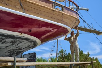 A worker works on the red hull of a wooden sailing ship in the dry dock, Patmos Marine, boatyard,