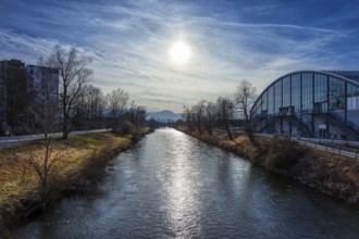 A river under a bridge in an urban area, surrounded by bare trees and a modern building in the