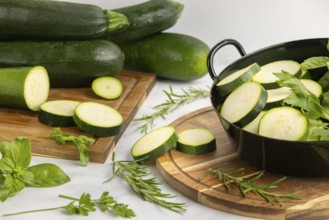 Sliced courgettes and fresh herbs in a black pot next to stacked courgettes on a wooden board