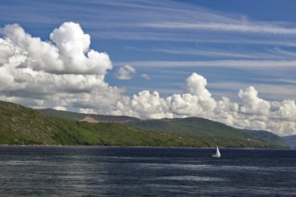 Single sailing boat on the sea, surrounded by a mountain landscape, under a sky with many clouds,