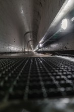 Long concrete tunnel with grid floor and damp surfaces, illuminated by lamps on the sides, Rappbode