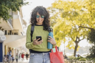 Smiling woman with curly hair walks through the city at sunset, holding a water bottle and red bag.