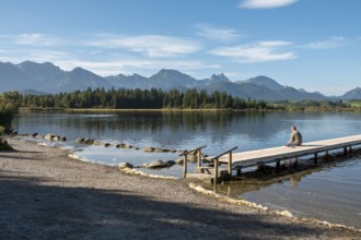 Person sitting on a wooden footbridge looking at a calm lake with mountains in the background,