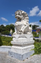 A stone lion statue on a pedestal, surrounded by plants under a blue sky with clouds, Bacalhôa,