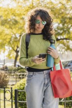 Woman with curly hair and sunglasses uses her smartphone in a park at sunset. Dressed casually in a