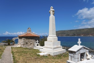 A tall monument next to a chapel in a cemetery with a view of the sea and surrounding mountains,