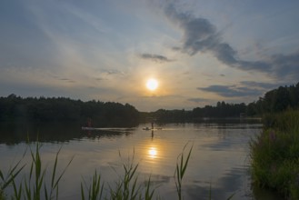 Calm lake at sunset, surrounded by trees and reeds, with a clear sky and scattered clouds, Oleš