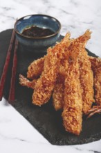 Shrimp in tempura, deep fried, on black slate, with sauce, light marble table, no people