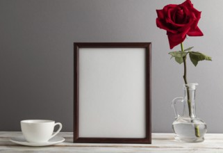 Brown wooden frame mockup with red rose in glass and white coffee cup on gray paper background.