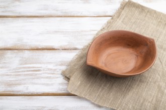 Empty clay brown bowl on white wooden background and linen textile. Side view, close up, copy space