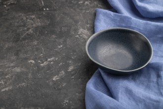 Empty blue ceramic bowl on black concrete background and blue linen textile. Side view, copy space