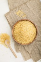 Wooden bowl with raw golden rice and wooden spoon on a white wooden background and linen textile.