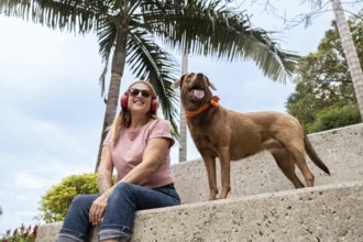 A smiling woman listens to music while sitting next to her alert dog wearing a colorful bandana