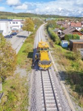 Yellow vehicle on ballast track in residential area under blue sky with clouds, tamping machine,