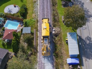 Yellow train runs on rails through a village, surrounded by houses and gardens, tamping machine,