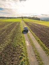 Black car driving on a narrow country lane through wide fields under a cloudy sky, Carsharing, VW