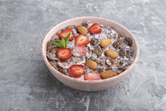 Chocolate cornflakes with milk, strawberry and almonds in ceramic bowl on gray concrete background.