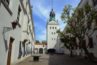 A sunny street leads to a historic building with a distinctive tower under a blue sky with clouds,