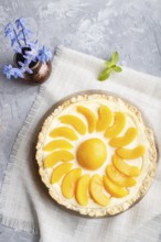 Round peach cheesecake and ceramic vase with blue flowers on a linen napkin on a gray concrete