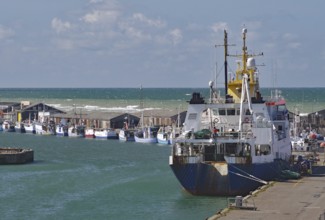 A large fishing boat moored at the harbour, with small fishing boats in the distance and the sea in