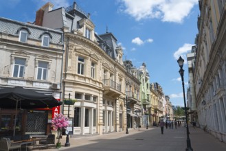 A lively city street with historic buildings under a blue sky on a sunny day, pedestrianised