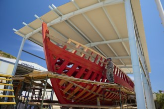 A partially built red wooden ship in a covered shipyard with a worker and blue sky, Patmos Marine,