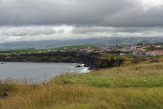 A village on the coast with green cliffs and a cloudy sky, Fenais da Luz, Sao Miguel Island,