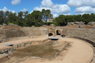 Partial view of a Roman amphitheatre with sandy floor and surrounding trees under a blue sky,