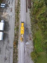 A yellow construction machine on a railway track next to a storage yard and some vehicles, tamping