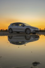 Silver car reflected in a puddle of water at sunset, atmospheric sky in shades of yellow and