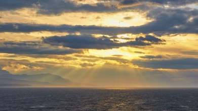 The sun sends its rays through the clouds over the sea near a mountain, Cape Melagkavi, Gulf of