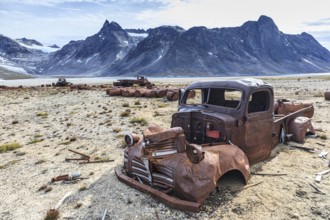 Rusty cars and oil drums in front of steep mountains, remains of a US airbase from the Second World