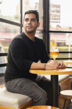 Handsome young Latino man sitting in a cafe with a glass of beer, gazing out the window. The cozy