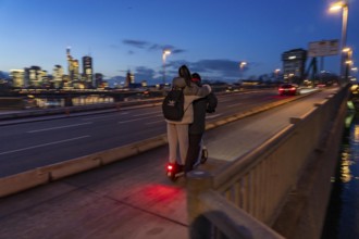 Skyline of the city centre of Frankfurt am Main, e-scooter, 2 persons, on the raft bridge, dusk,