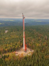 A crane lifts parts of a wind turbine in an autumnal forest, wind farm construction site, Grömbach,