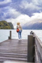 Person standing on a wooden footbridge with a view of the lake and mountains under a cloudy sky,