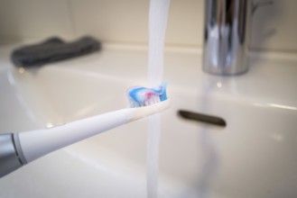 A bathroom tap is running while a toothbrush is held under the running water, symbolic image of