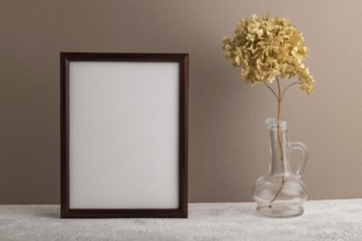 Brown wooden frame mockup with dried hydrangea in glass on beige paper background. Blank, vertical