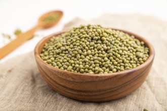 Wooden bowl with raw green mung bean and wooden spoon on a white background and linen textile. Side