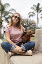 Middle-aged woman with sunglasses, sitting on a cement bench next to her dog, with palm trees in