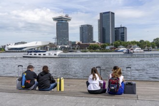 Tourists sitting at the back of Amsterdam Centraal railway station, on the promenade of the river