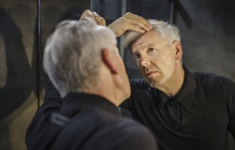 Symbolic photo on the subject of a high hairline and receding hairline. A man looks at his hairline