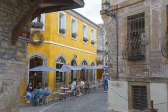 Busy people sitting in front of historic buildings under parasols in an old town alley, Xanthi,